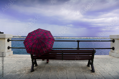 An unrecognisable woman holding an umbrella with a red and black squared pattern is sitting on a wooden bench, looking at a rough sea during a storm in Bari, Italy.