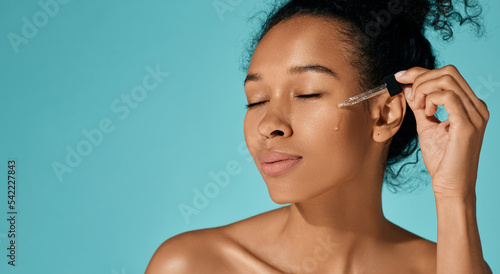 Facial skin care with essential oil and serum. Happy African woman applying essential oil on her face for hydrated skin with pipette dropper, isolated on blue background photo