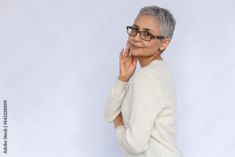 Portrait of confident senior woman posing over white background. Mature Caucasian woman wearing eyeglasses and white jumper touching face and looking at camera. Aging woman beauty concept Portrait of confident senior woman posing over white background. Mature Caucasian woman wearing eyeglasses and white jumper touching face and looking at camera. Aging woman beauty concept