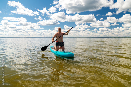 A man kneeling on a SUP board with a paddle in the lake on a sunny day against the backdrop of white clouds.