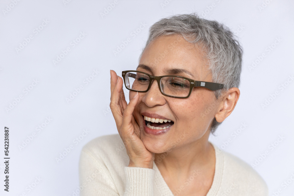 Fototapeta premium Close-up of senior woman laughing and touching face over white background. Mature Caucasian woman wearing eyeglasses and white jumper looking away and smiling. Happiness concept