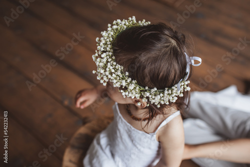 Portrait of pretty one year girl in white summer dress, flower crown and with eco beads sitting in the studio near mom's legs. Eco style concept