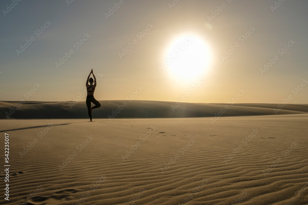 Mulher pratica pose de yoga sobre duna durante o por-do-sol.