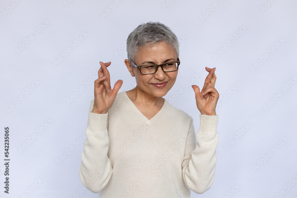 Portrait of deceitful senior woman making liar gesture. Mature Caucasian woman wearing eyeglasses and white jumper crossing fingers in hope over white background. Liar, hope, wish concept - obrazy, fototapety, plakaty 