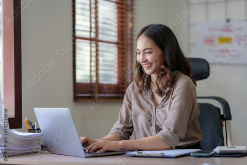 Happy Asian businesswoman sitting in the office with her laptop computer working diligently and smiling brightly with loads of papers on her desk.