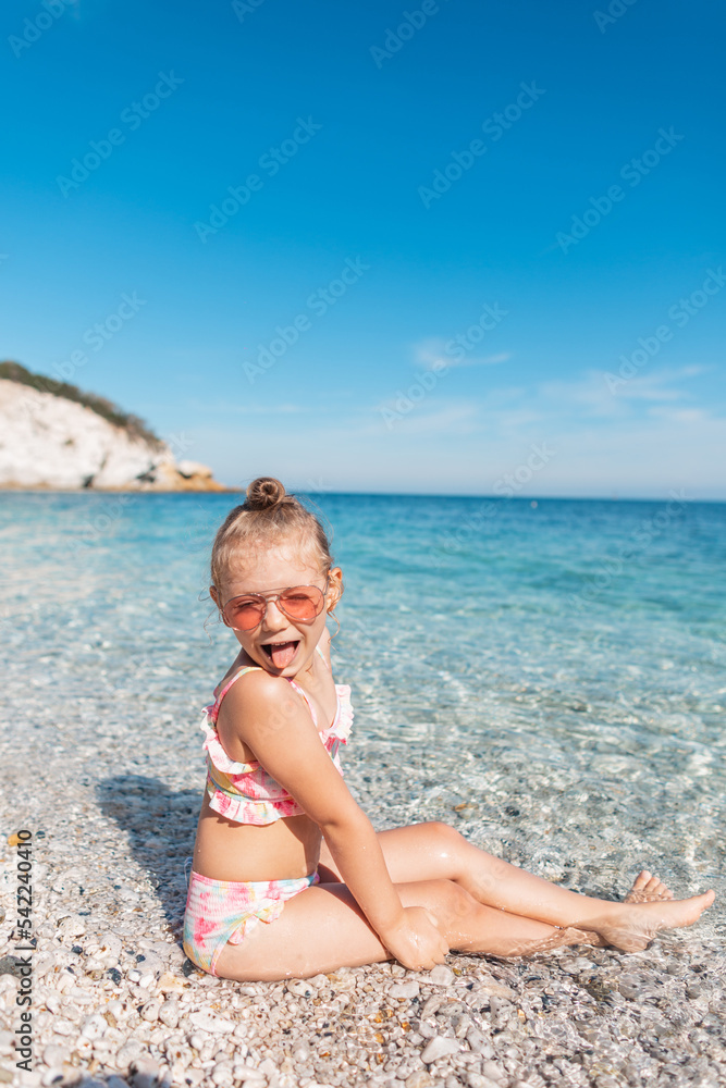 Funny beautiful happy baby girl with sunglasses in a fashion bathing suit is sitting and having fun on the beach by the clear blue sea. Summer holidays Funny beautiful happy baby girl with sunglasses in a fashion bathing suit is sitting and having fun on the beach by the clear blue sea. Summer holidays