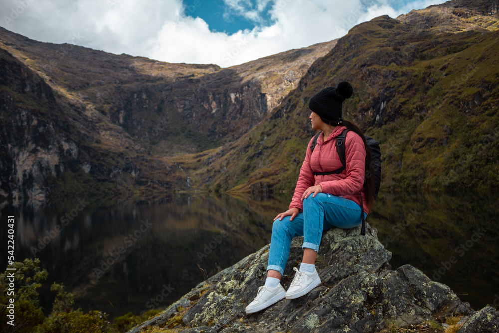 Mujer alegre con mochila sentada en una roca contra un lago rodeado de montañas