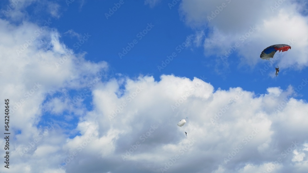 Two skydiver athletes are flying down on their parachutes against a beautiful blue sky with white clouds. View from the ground of two beautifully gliding skydivers. The concept of extreme sports. Two skydiver athletes are flying down on their parachutes against a beautiful blue sky with white clouds. View from the ground of two beautifully gliding skydivers. The concept of extreme sports.