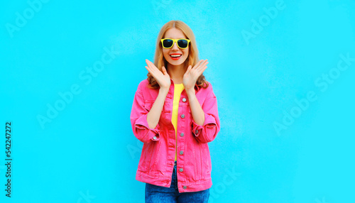Portrait of happy excited surprised young woman wearing colorful clothes, pink jacket, yellow sunglasses on blue background