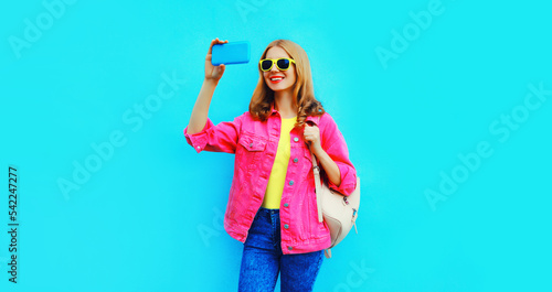 Portrait of happy smiling stylish young woman taking selfie with smartphone wearing pink jacket, backpack, yellow sunglasses on blue background