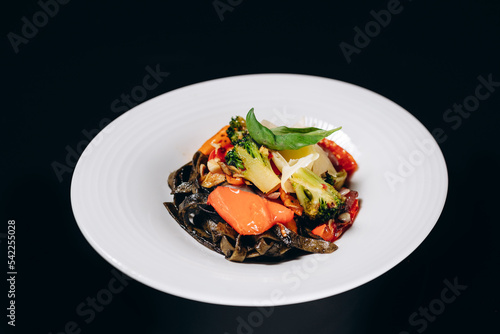 Vegetable pasta with cuttlefish ink, in a white plate on a black isolated background.
