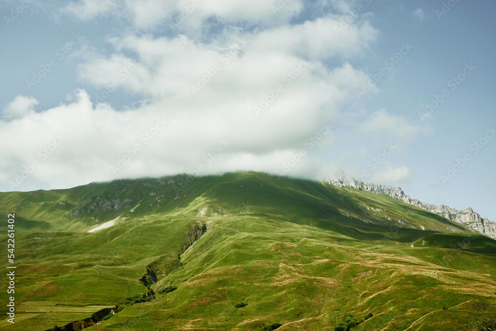 Mountain peaks covered with green meadows and clouds above them. Midday in mountains. North Ossetia-Alania, Russia. Mountain peaks covered with green meadows and clouds above them. Midday in mountains. North Ossetia-Alania, Russia.