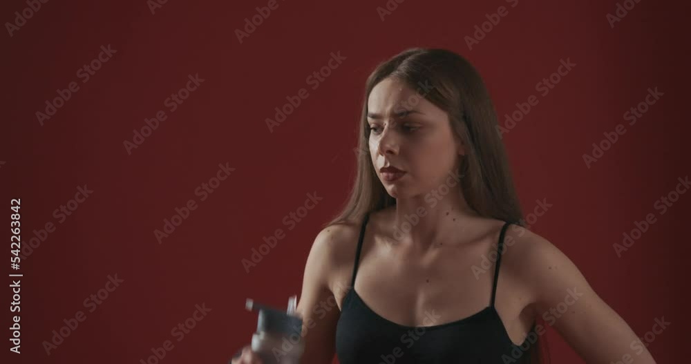 Side view of slim, fit girl with long hair working out indoors, standing, holding bottle, water drinking. Pretty young woman bodybuilding. Isolated on red studio background.