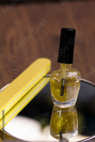 A bottle of translucent nail polish with gold glitter and a yellow nail file on a mirror surface