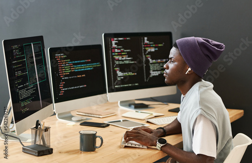 Young serious IT engineer in casualwear looking at coded data on computer screens while typing on keyboard by workplace