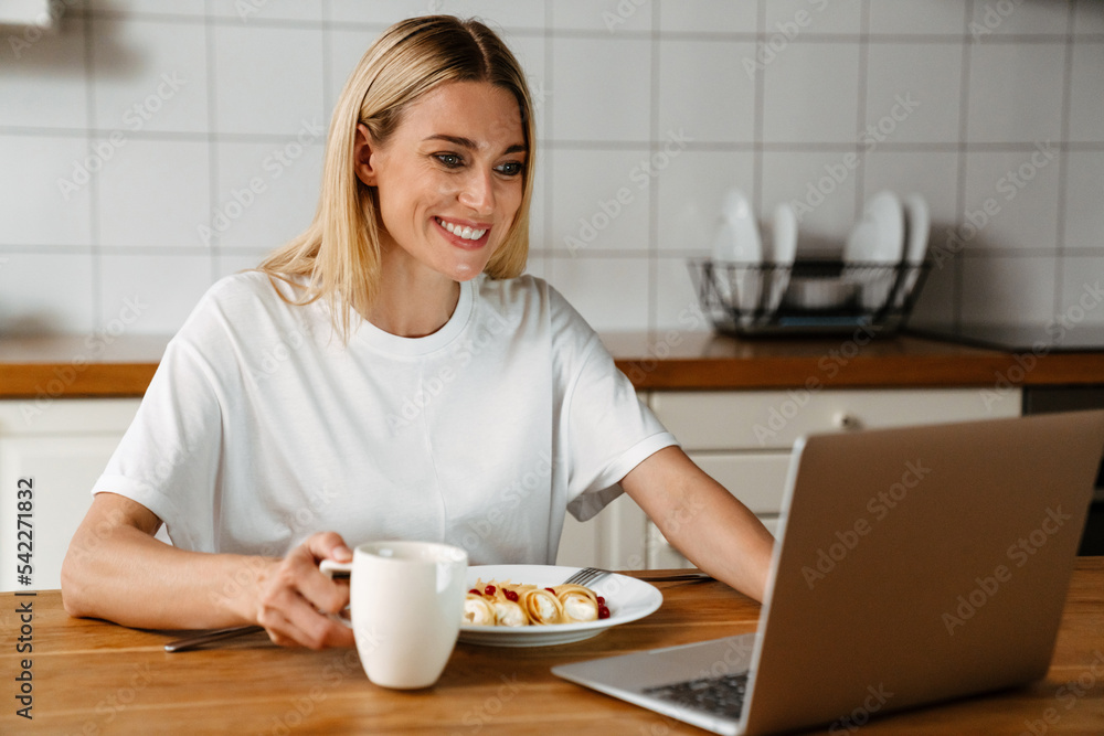 Happy woman smiling and using laptop while having breakfast at home