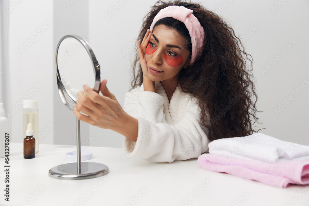Happy tanned pretty curly Latin lady with pink patch for eye looks at mirror doing beauty procedure in white home interior. Copy space. Moisturizing beauty grooming, skincare and pampering concept Happy tanned pretty curly Latin lady with pink patch for eye looks at mirror doing beauty procedure in white home interior. Copy space. Moisturizing beauty grooming, skincare and pampering concept