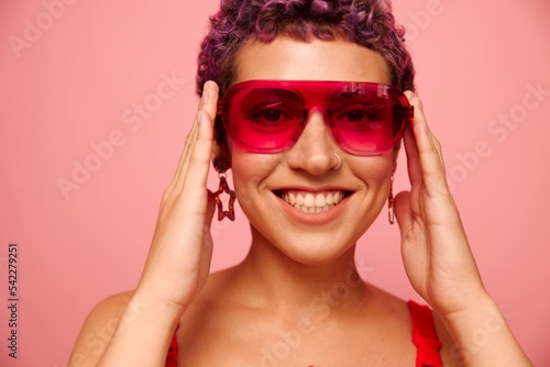 Fashion portrait of a woman with a short haircut in colored sunglasses and a smile with teeth in a red top on a pink background