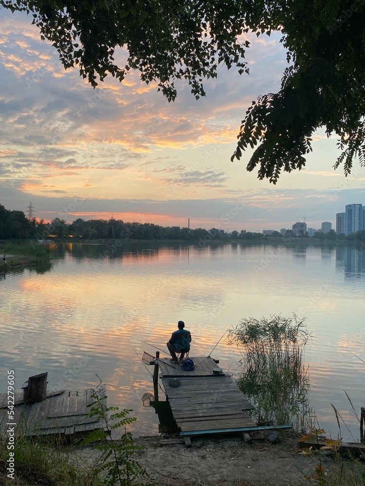 Silhouette of a fisherman who catches fish on a fishing rod on the shore of a beautiful lake with reeds. There's a sunset on the horizon. The sun is reflected in the water Silhouette of a fisherman who catches fish on a fishing rod on the shore of a beautiful lake with reeds. There's a sunset on the horizon. The sun is reflected in the water