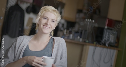 Medium shot of attractive young woman with short blonde hair, smiling and drinking coffee from a white mug in an urban loft