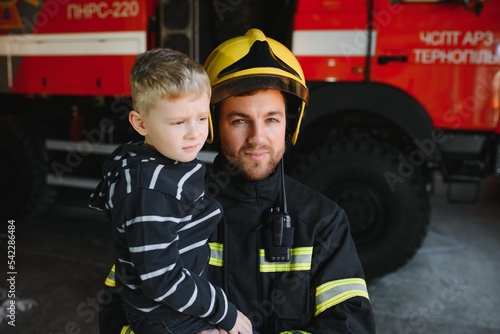 Dirty firefighter in uniform holding little saved boy standing on black background.