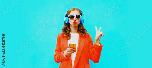 Portrait of stylish young woman in headphones listening to music with smartphone and blowing her lips on blue background