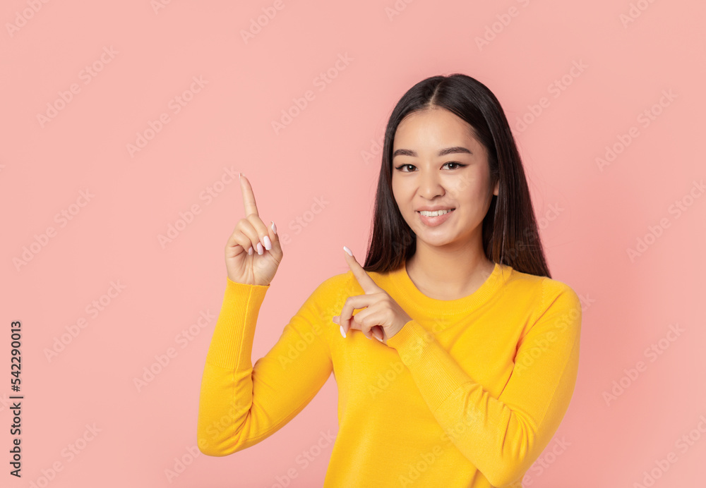 Beautiful Asian young woman smiling with his finger pointing on a pink background with copy space, looks at the camera and smiles friendly Beautiful Asian young woman smiling with his finger pointing on a pink background with copy space, looks at the camera and smiles friendly