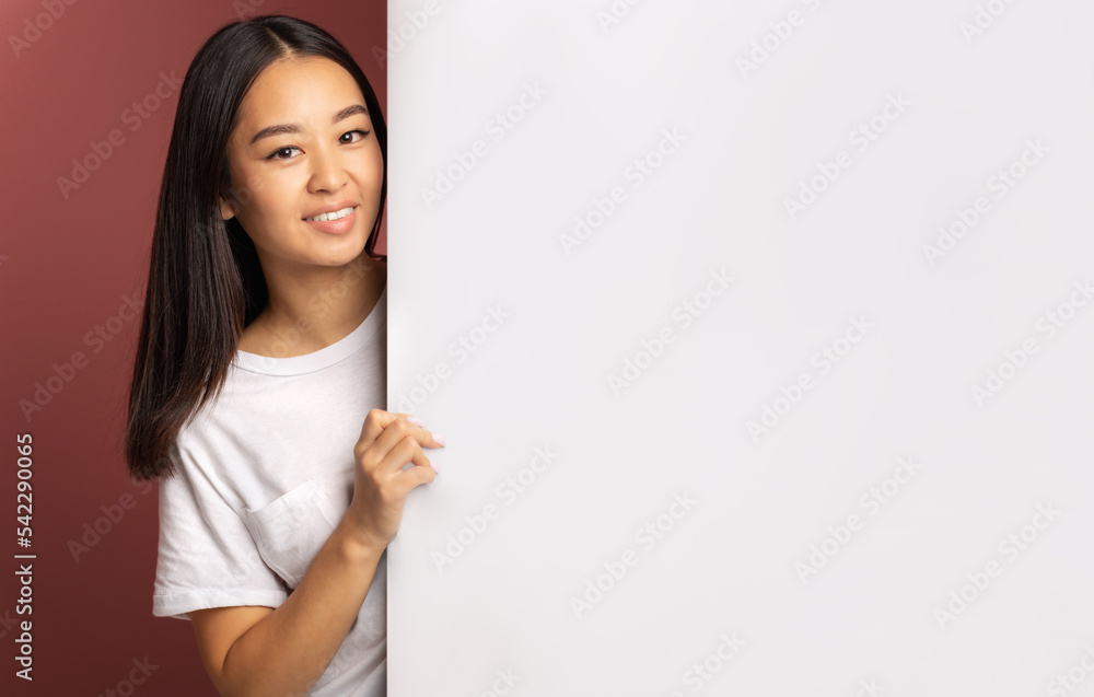 Happy smiling Asian woman peeking out from empty white board, looking at the camera and smiles Happy smiling Asian woman peeking out from empty white board, looking at the camera and smiles