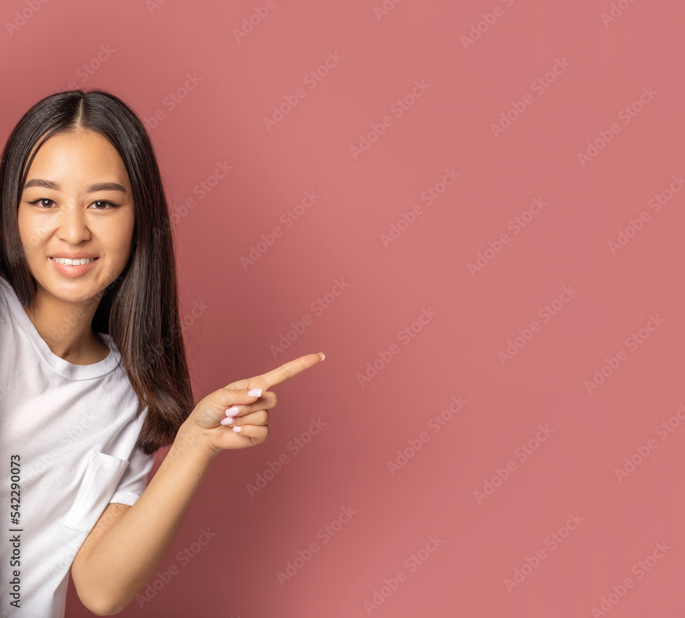 Beautiful Asian young woman smiling with his finger pointing on a pink background with copy space, looks at the camera and smiles friendly