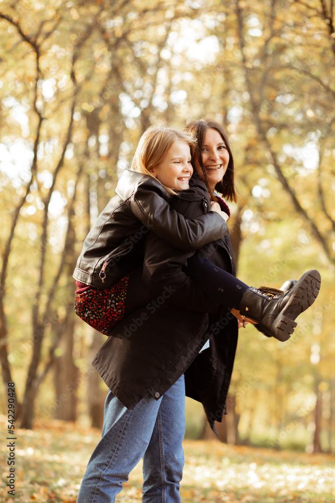 Rejoicing young woman ride on back little girl side view. Happy mother and daughter in casual black outfits have fun and play in golden autumn park. Fall nature, active lifestyle, family weekend