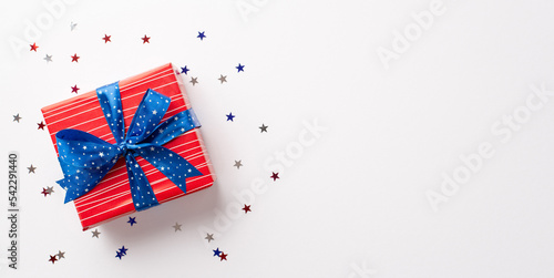 Forth of July concept. Top view photo of red giftbox with blue ribbon bow and star shaped confetti on isolated white background with empty space