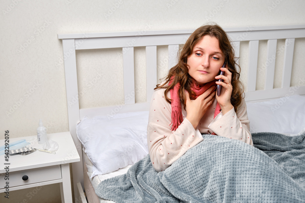 A sick woman is holding a sore throat with a phone in her hand, neck pain. Adult ill woman sitting on a home bed in a white bedroom, female aged 35 years A sick woman is holding a sore throat with a phone in her hand, neck pain. Adult ill woman sitting on a home bed in a white bedroom, female aged 35 years