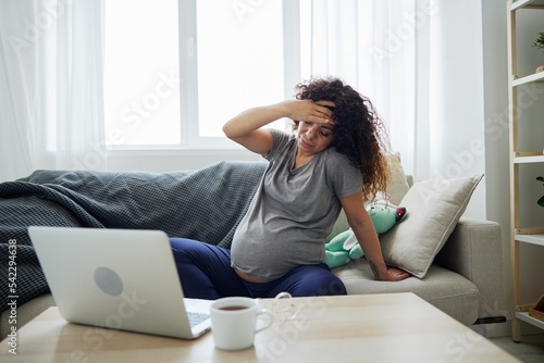 Pregnant woman with headache and fatigue sits at home on sofa with a laptop and watches tv shows movies and series. Lifestyle of a pregnant woman, preparation for childbirth, last month of pregnancy © SHOTPRIME STUDIO