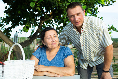 Portrait of positive man hugging his senior mother who sitting at table under tree in garden. © JackF