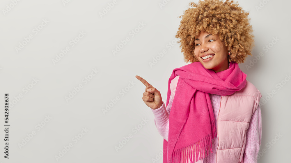 Fashionable curly haired woman in vest and pink warm scarf points index finger left shows promo offer demonstrates banner smiles gladfully isolated over grey background. Follow this direction