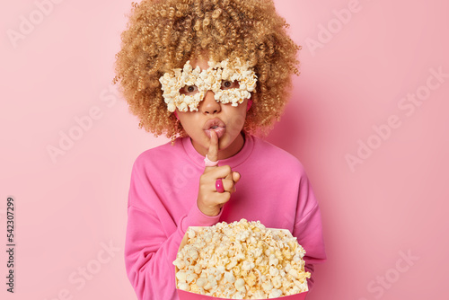 Mysterious curly woman makes silence gesture presses index finger to lips shares secret poses with tasty salty popcorn focused directly at camera wears pullover and eyeglasses isolated on pink wall
