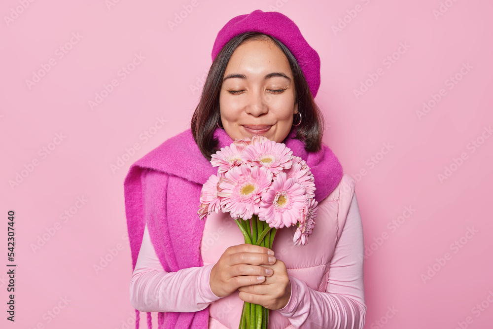 Delighted Asian woman enjoys fragrant from gerberas holds bouquet likes flowers keeps eyes closed wears beret and scarf has date with boyfriend isolated over pink background. Feminine concept Delighted Asian woman enjoys fragrant from gerberas holds bouquet likes flowers keeps eyes closed wears beret and scarf has date with boyfriend isolated over pink background. Feminine concept