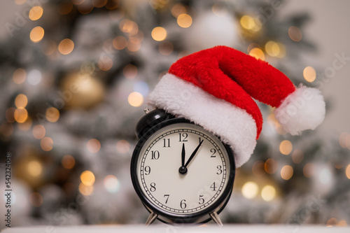 Alarm clock in a santa hat against the background of a christmas tree.