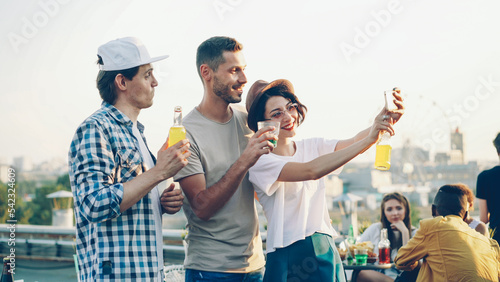 Pretty young woman is using smartphone to take selfie with her cheerful male friends, happy young people are posing then laughing
