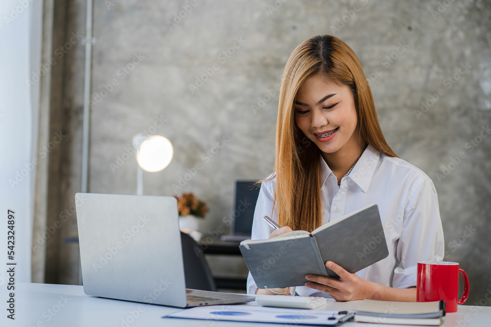 Asian woman working in financial accounting, analyzing charts using calculator, laptop with documents and holding pens, sits in a chair at a desk in a financial office. Asian woman working in financial accounting, analyzing charts using calculator, laptop with documents and holding pens, sits in a chair at a desk in a financial office.