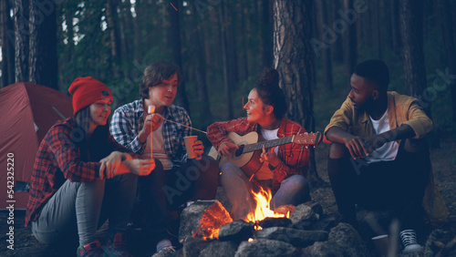 Pretty young woman is playing the guitar while her friends are singing funny songs and laughing cooking marshmallow on fire and clapping hands. Music, nature and fun concept.