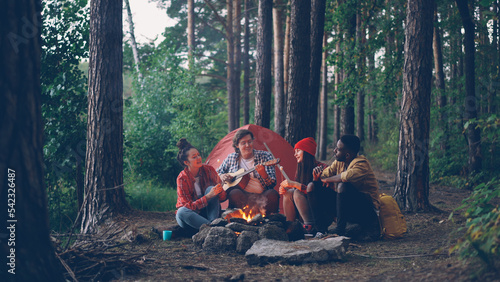 Man tourist in casual clothing is playing the guitar while his friends are cooking and eating sweet marshmallow sitting around campfire on autumn day. People and camp fun concept.