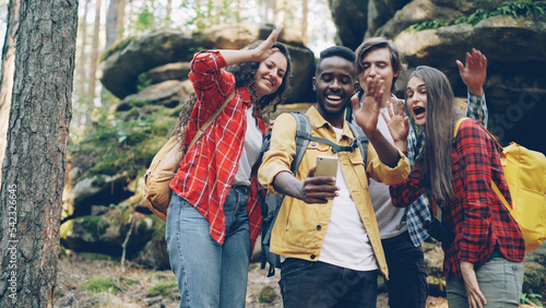 Happy African American guy is making video call using modern smartphone during hike in forest with friends, young people are looking at screen, waving hand and talking.