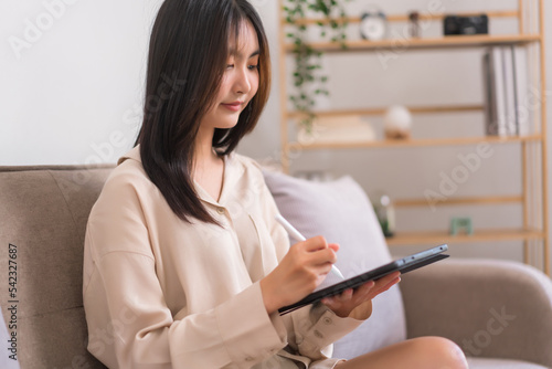 Lifestyle at home concept, Young Asian woman writing on tablet to working while sitting on sofa © Pichsakul