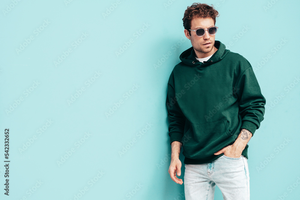 Handsome confident hipster model. Sexy unshaven man dressed in summer stylish green hoodie and jeans clothes. Fashion male with curly hairstyle posing in studio. Isolated on blue. In sunglasses Handsome confident hipster model. Sexy unshaven man dressed in summer stylish green hoodie and jeans clothes. Fashion male with curly hairstyle posing in studio. Isolated on blue. In sunglasses