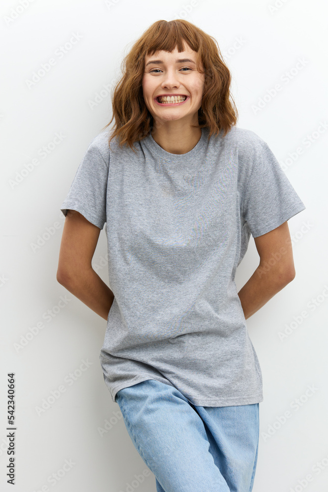 Obraz premium a wide-smiling, joyful woman stands in a gray T-shirt on a white background, leaning against the wall. Vertical Studio Photography