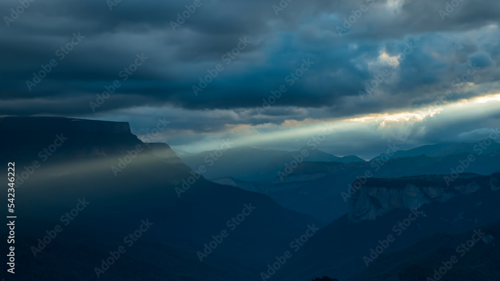 Rain-clouds over beautiful mountain gorge. The slanting rays of the evening sun break through them. A beautiful mysterious mountain landscape. Mountains and gorges in beautiful sunlight.