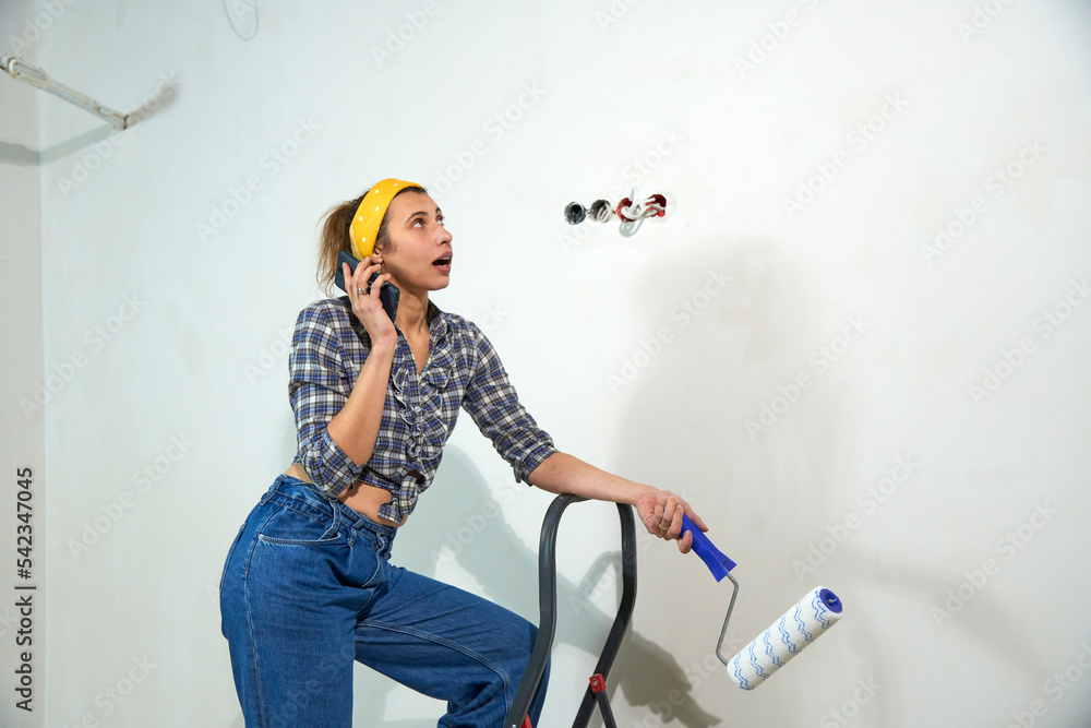 Young artist paints the walls in the apartment with a roller. Beautiful woman is working on a new bedroom project. Cute female in blue jeans and t-shirt speaking with cellphone. Renovations at home. Young artist paints the walls in the apartment with a roller. Beautiful woman is working on a new bedroom project. Cute female in blue jeans and t-shirt speaking with cellphone. Renovations at home.