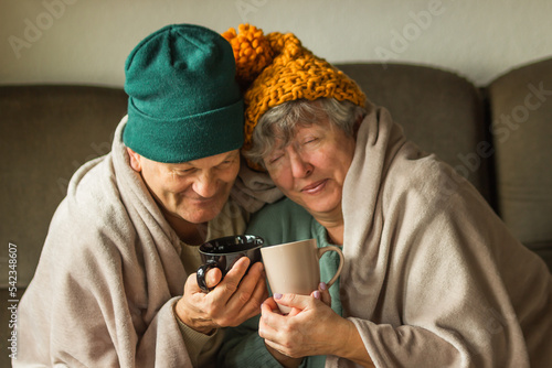 Grandparents in knitted caps wrapped in warm blanket hold cups of hot tea or coffee, feeling cold at home. Happy couple.