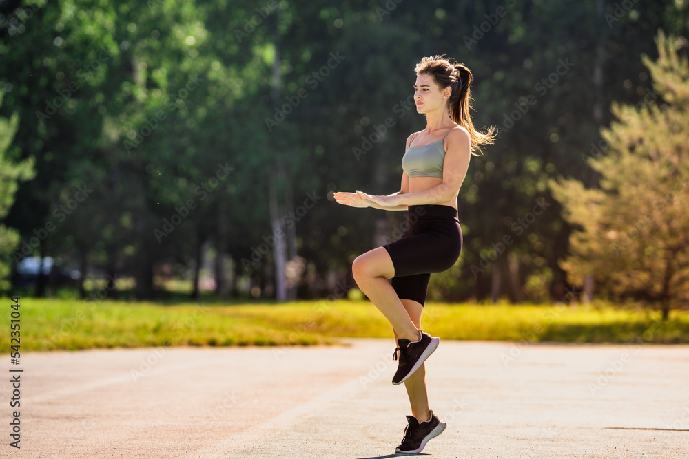 Fit hispanic brunette girl dressed in sportswear making workout outdoor jumping agains blurry park on background. Beautiful Italian girl practicing sport at park. Young woman makes exercises outdoor.